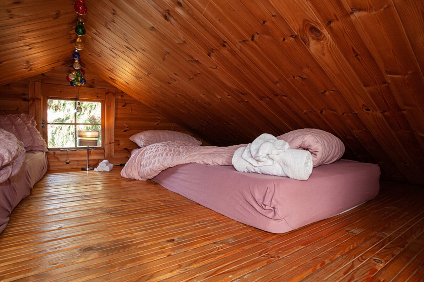 Un dormitorio acogedor en el ático con suelos de madera, una cama rosa y una ventana con vistas al verdor.