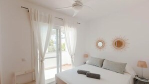 Une chambre à coucher avec un lit, deux miroirs décoratifs en forme de soleil fixés au mur, un ventilateur de plafond et une fenêtre ornée de rideaux légers.