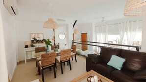 Un salon moderne avec un canapé marron foncé, des fauteuils en osier, une table à manger en verre et un balcon agrémenté de rideaux blancs.