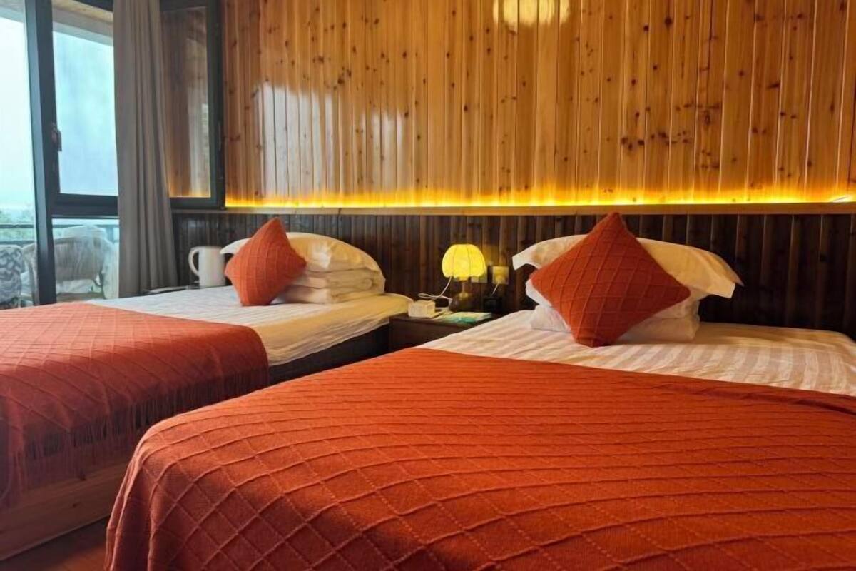 Two beds with red bedding and white pillows, a wooden wall with vertical slats, a window with curtains, and a nightstand with a lamp.
