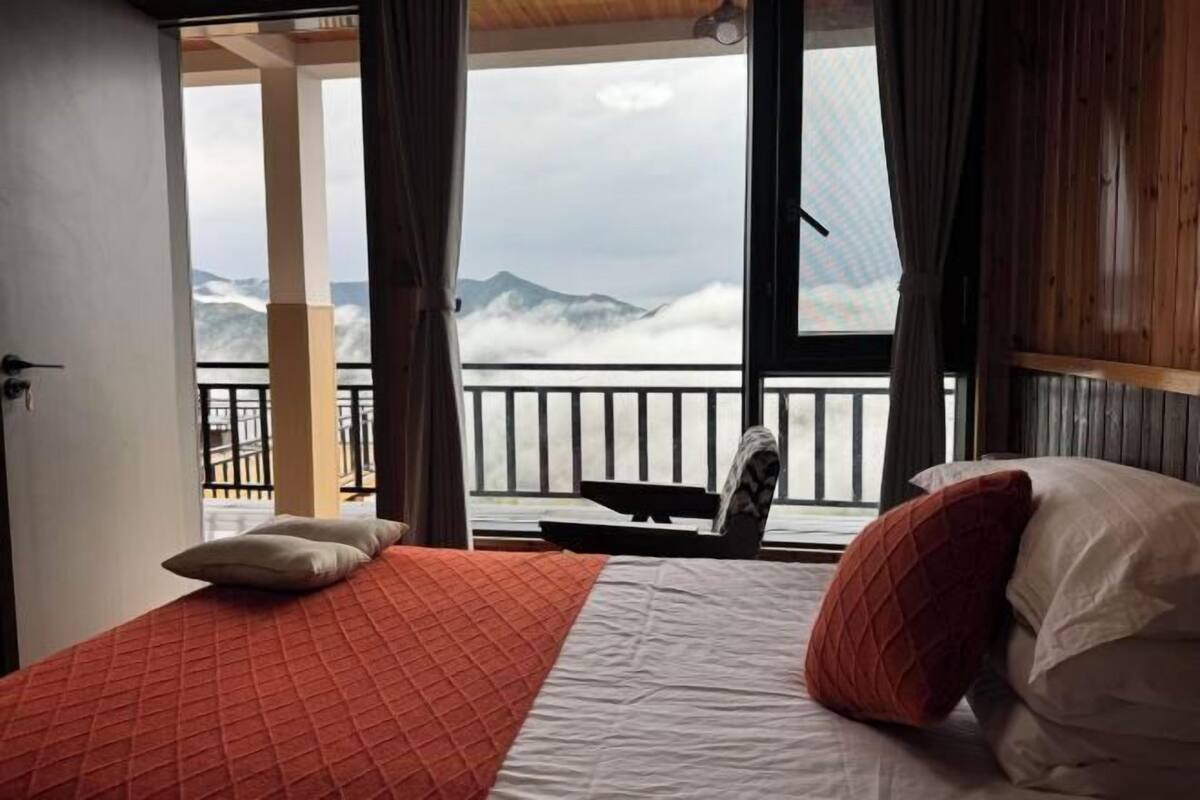 A bedroom with a bed, pillows, a chair, and a view of mountains through a balcony.