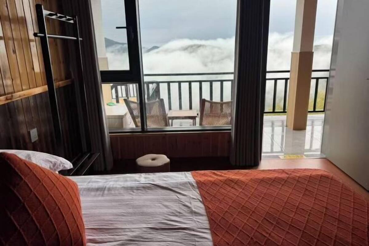 A bedroom with a bed, a small stool, a window with a view of mountains, and a balcony with chairs.