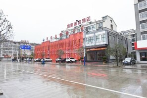 A building with a red facade and signage, a parking lot with cars, and a wet ground indicating recent rain. - Fenghuag Xidi Hotel (Jinzhai)