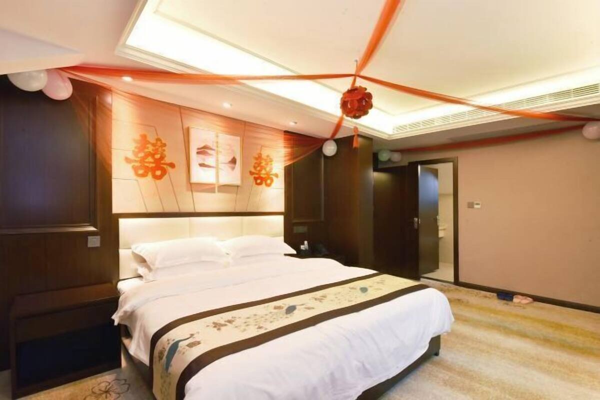 A hotel room with a large bed, a headboard with artwork, a ceiling with a light fixture, and a door.