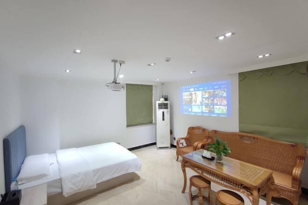 A hotel room with a single bed, a dining table with chairs, a sofa, a ceiling fan, and a projector screen displaying images.