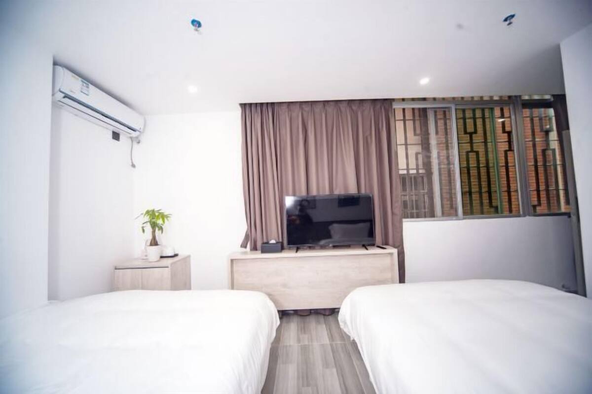 A modern hotel room with two single beds, a TV on a wooden dresser, a potted plant, and a window with curtains.