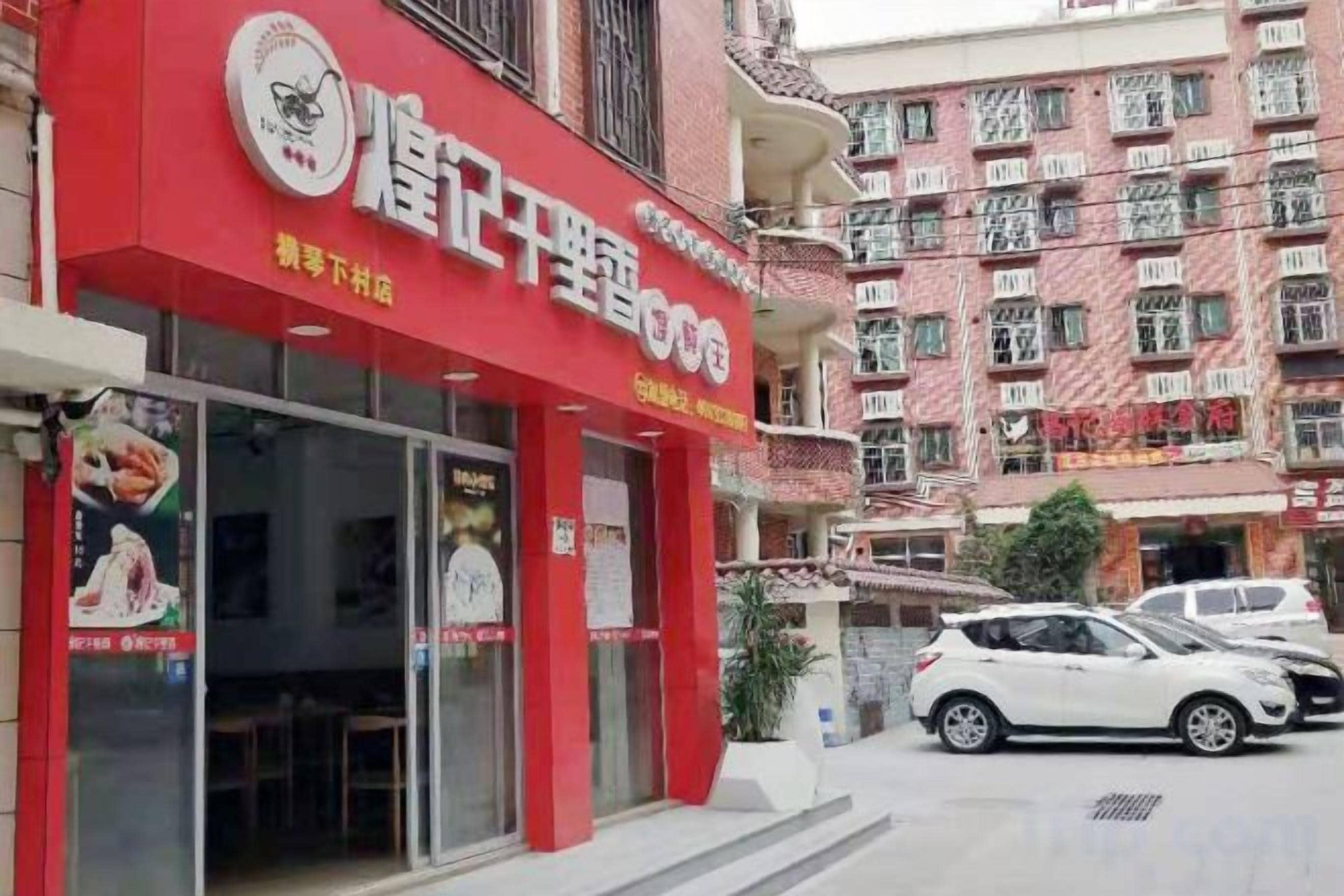 A red storefront with a sign, parked cars, and a multi-story building in the background.