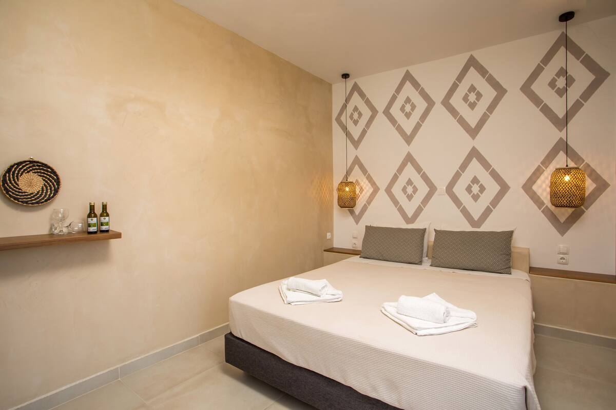 A hotel room with a large bed, two pillows, a bedside table with a bottle and glasses, and a wall with a geometric pattern.