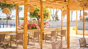 An outdoor seating area with wooden tables and chairs, hanging woven light fixtures, and a view of the beach and palm trees. - ATMA BEACH ROOMS & SUITES (Rhodes)