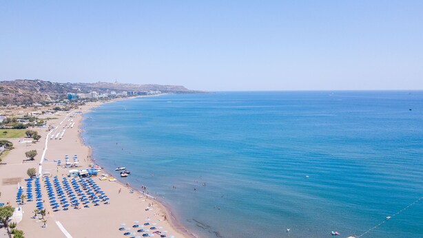 A coastal beach with rows of sun umbrellas and beach chairs, a sandy shoreline, and a clear blue sea. - ATMA BEACH ROOMS & SUITES (Rhodes)