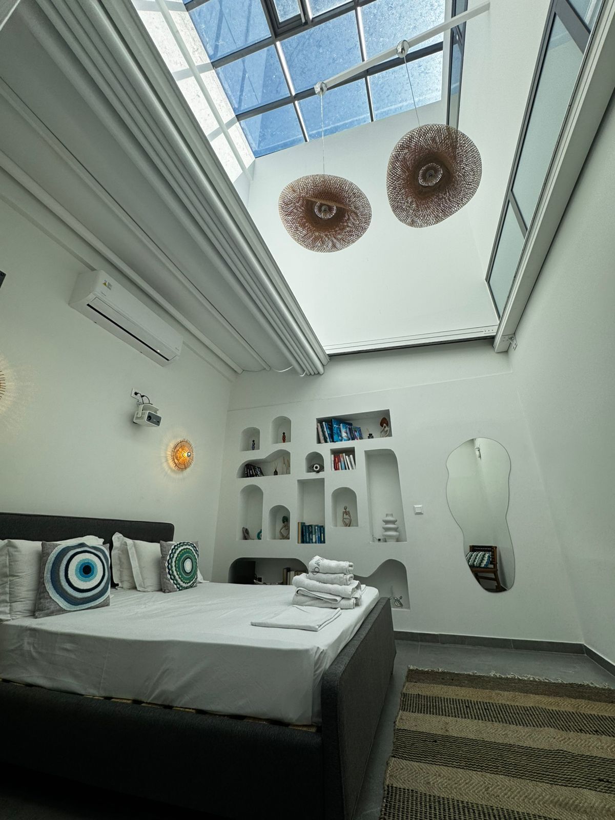 Ein modernes Schlafzimmer mit Oberlicht, einem Bett mit Kissen, einem Nachttisch, einem Spiegel und eingebauten Regalen mit Büchern und Dekoration.