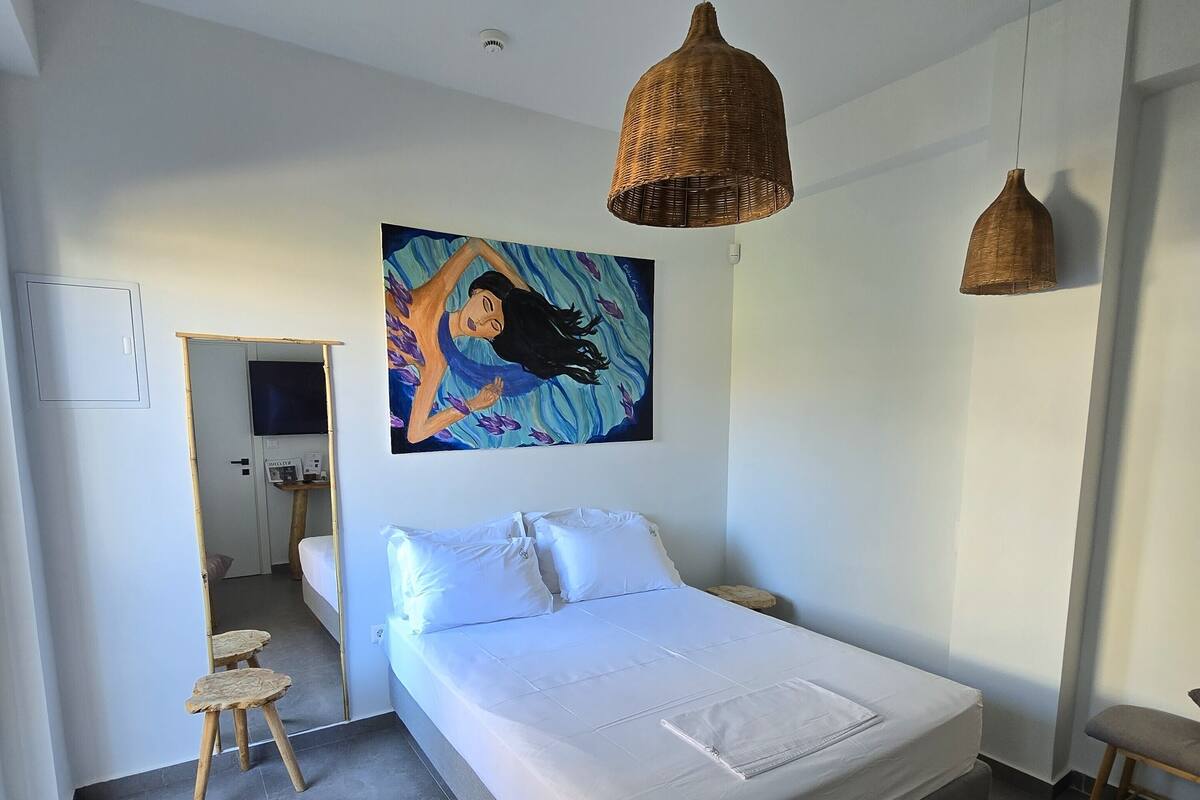 Ein modernes Schlafzimmer mit einem großen Bett, einem kleinen Hocker, einer Korbtrophäe als Pendelleuchte und einem Bild an der Wand.