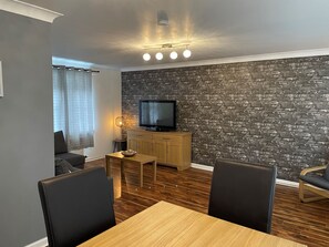 A modern living room with a brick wall, a flat-screen TV, wooden furniture, and a dining area. - Beautiful 2-bed Apartment in Renfrew (Renfrew)