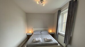 Ein ordentlich eingerichtetes Schlafzimmer mit einem großen Bett, zwei Nachttischen mit Lampen, einem Fenster mit Vorhängen und einer Deckenleuchte.