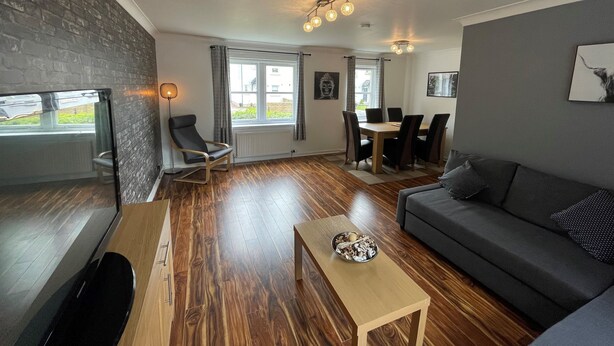Ein modernes Wohnzimmer mit Holzboden, einer grauen Couch, einem Holztisch, einem Fernseher, einem Essbereich mit Tisch und Stühlen sowie einer Ziegelwand.