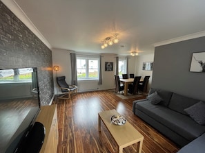 A modern living room with a wooden floor, a grey sofa, a wooden coffee table, a TV, a dining area with a table and chairs, and a brick wall. - Beautiful 2-bed Apartment in Renfrew (Renfrew)
