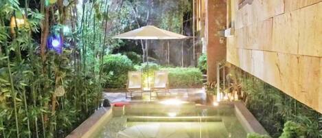 A serene indoor pool area with a clear water surface, surrounded by lush greenery and a seating area with a table and chairs.