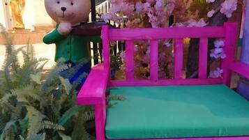 Un banco rosa y verde con una escultura de un osito de peluche, rodeado de flores rosadas y follaje.