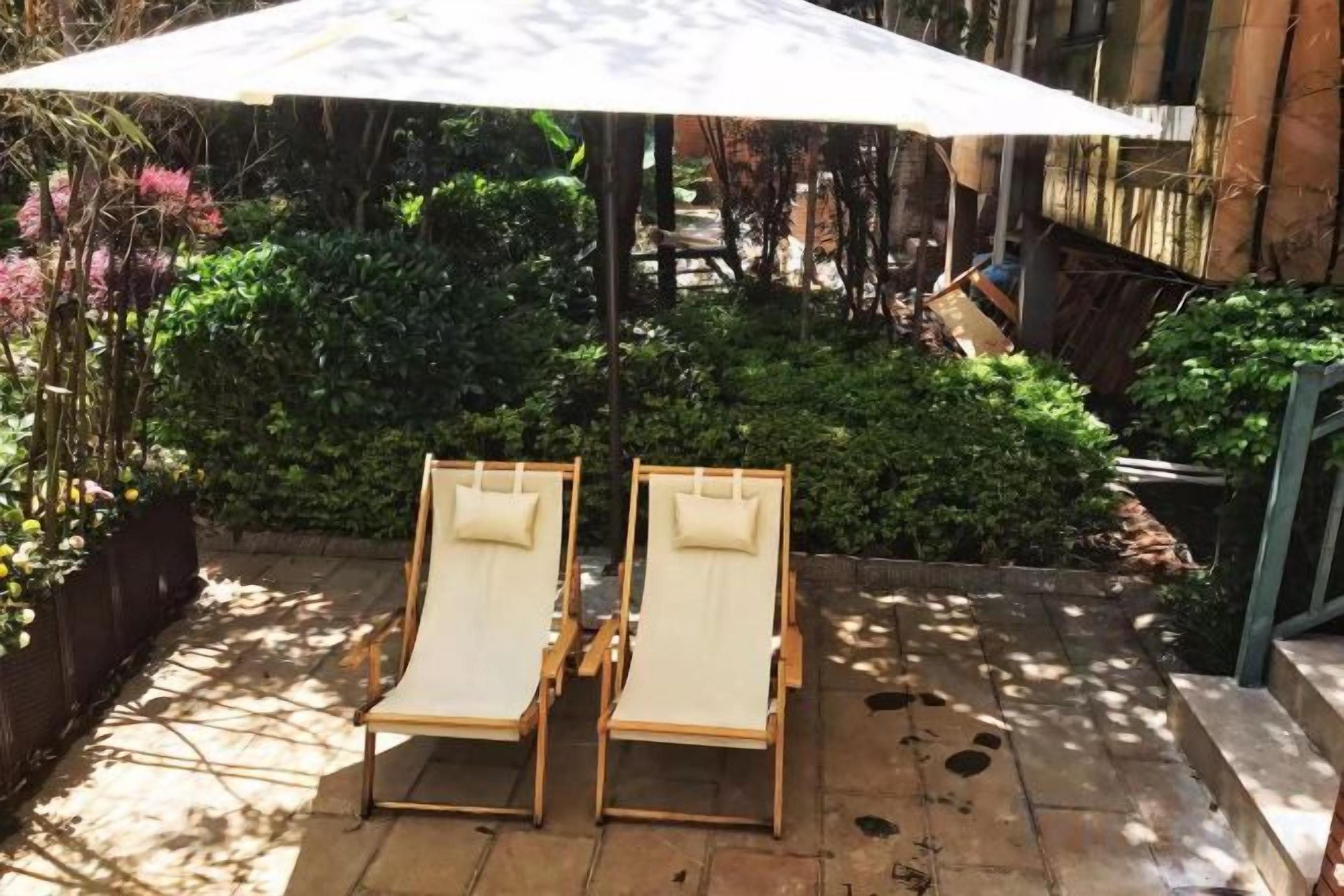 Two lounge chairs under a large umbrella in a garden area with greenery and a building in the background.