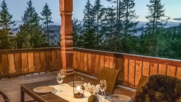 Un espace extérieur aménagé pour prendre les repas, avec une table dressée pour deux personnes, entouré d’une balustrade en bois et offrant une vue panoramique sur les montagnes en arrière-plan.