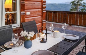 A dining table set with white plates, wine glasses, and a vase of white flowers on a wooden deck with a view of mountains.