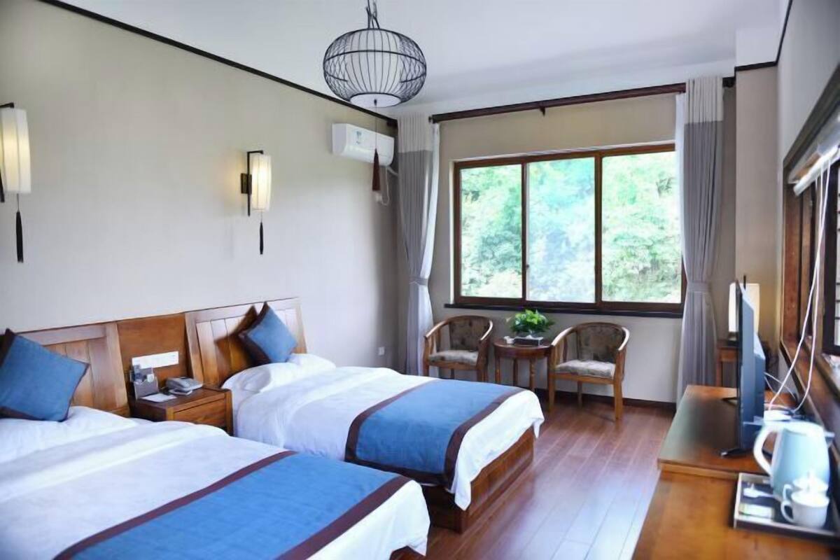 A hotel room with two beds, a small table with chairs, a TV, and a large window with curtains.