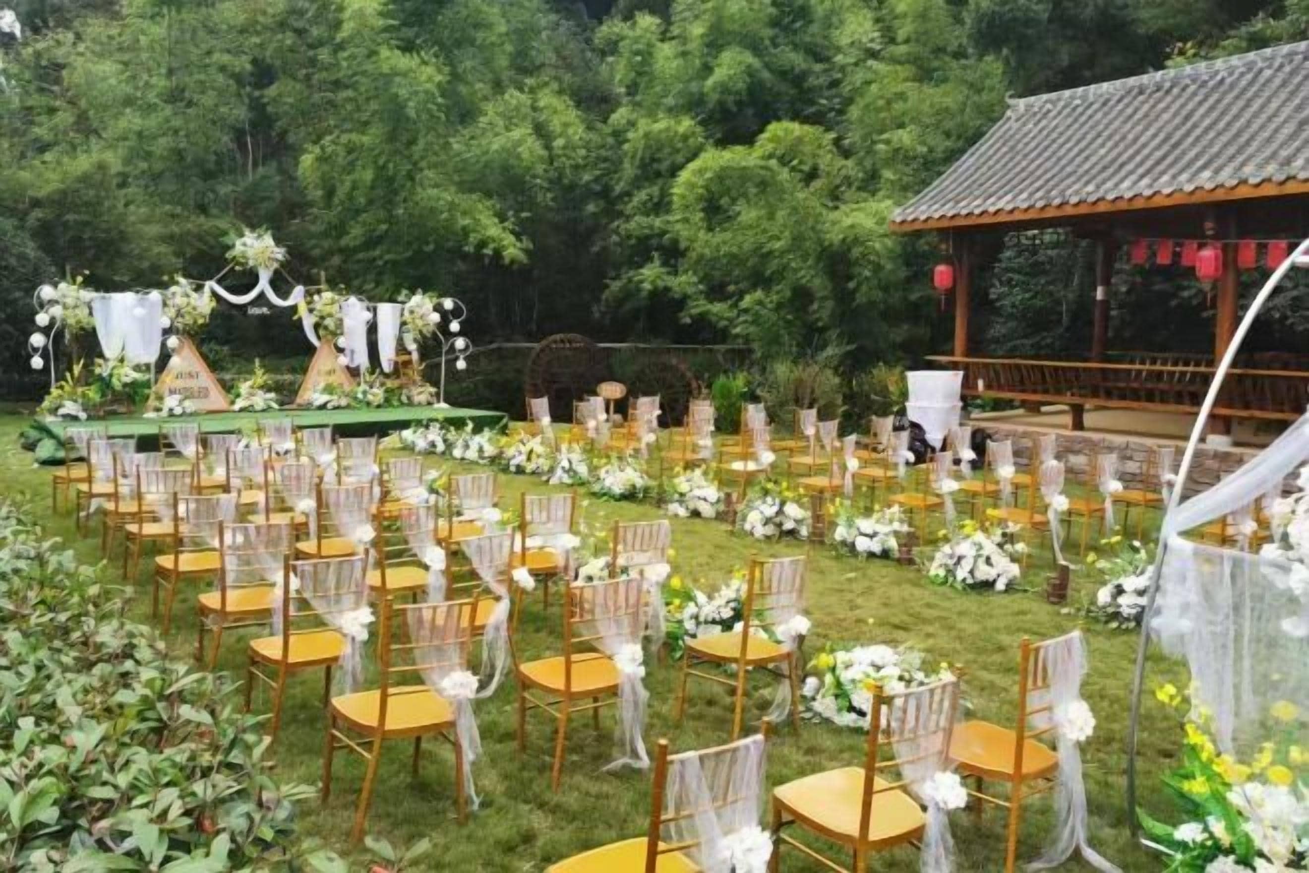 An outdoor wedding venue with rows of chairs and tables, a stage with floral decorations, and a gazebo in the background.