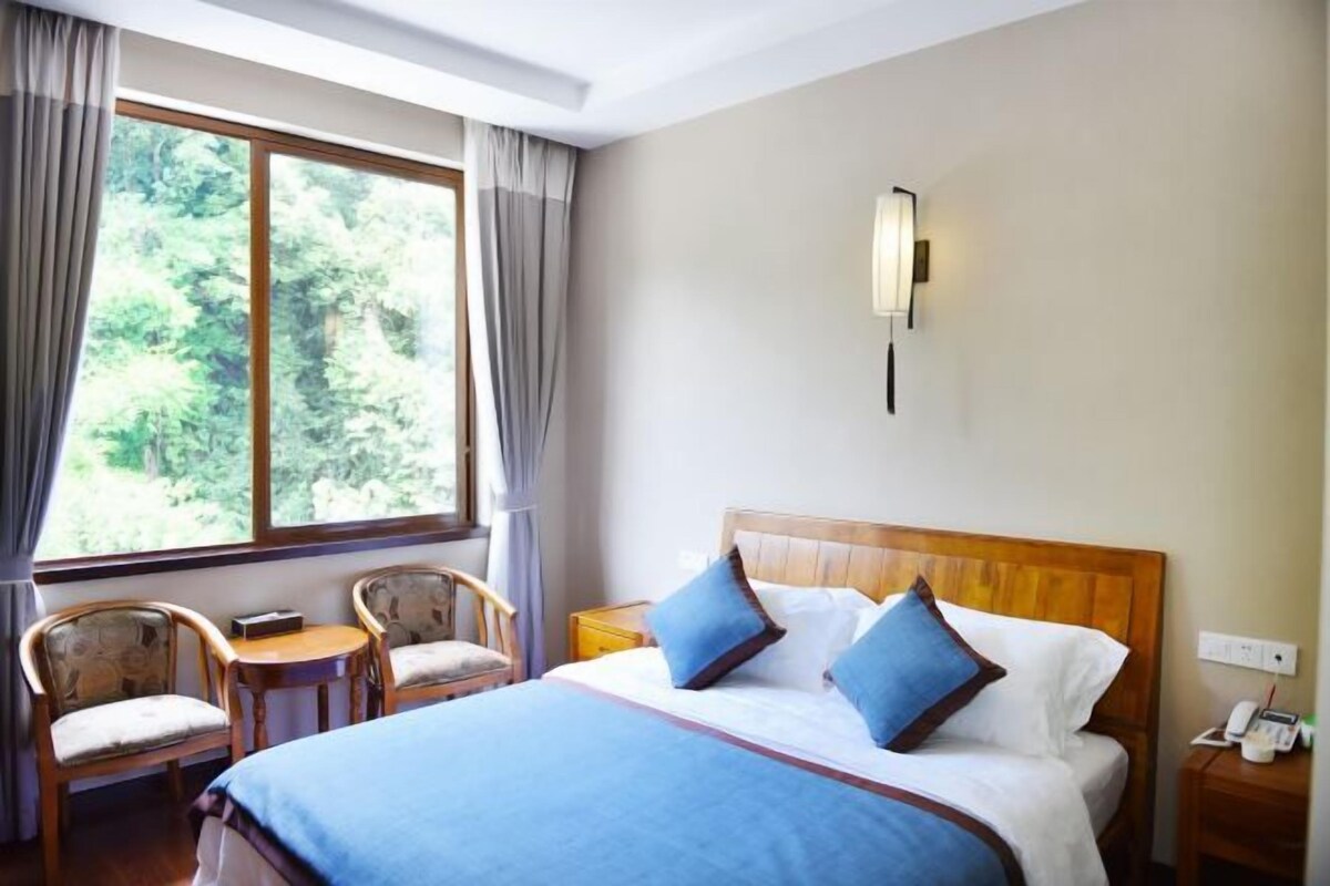 A hotel room with a large window, a bed with blue bedding, two chairs, a bedside table, and a wall-mounted light.