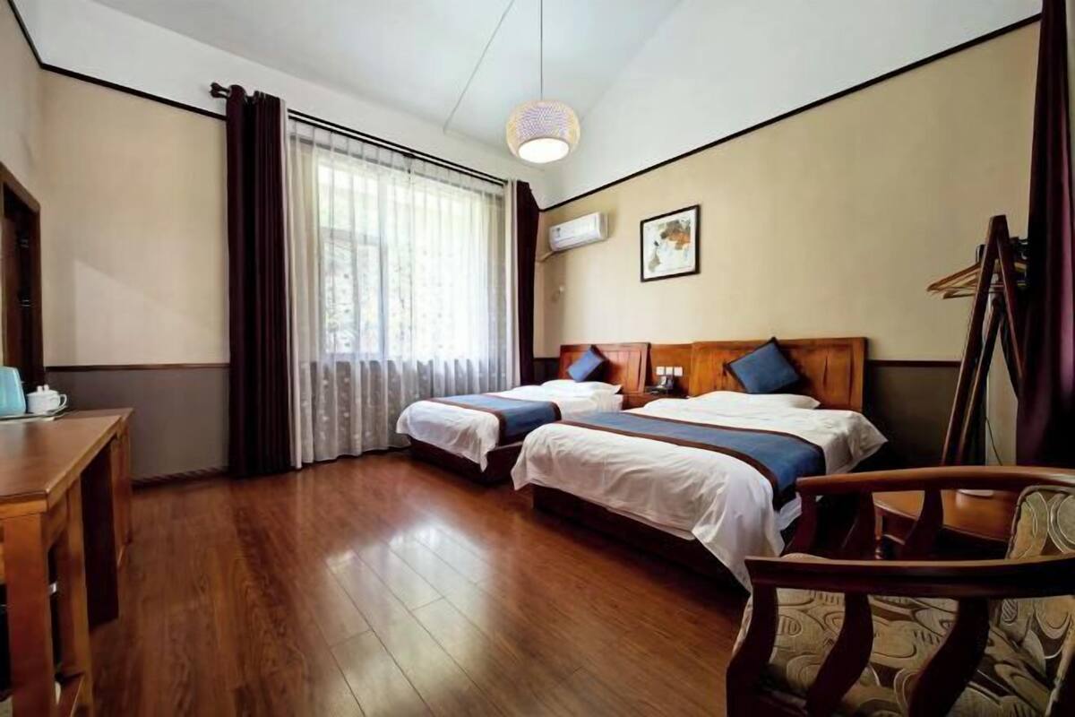 A hotel room with two beds, a wooden desk, a chair, a hanging light fixture, and a window with curtains.