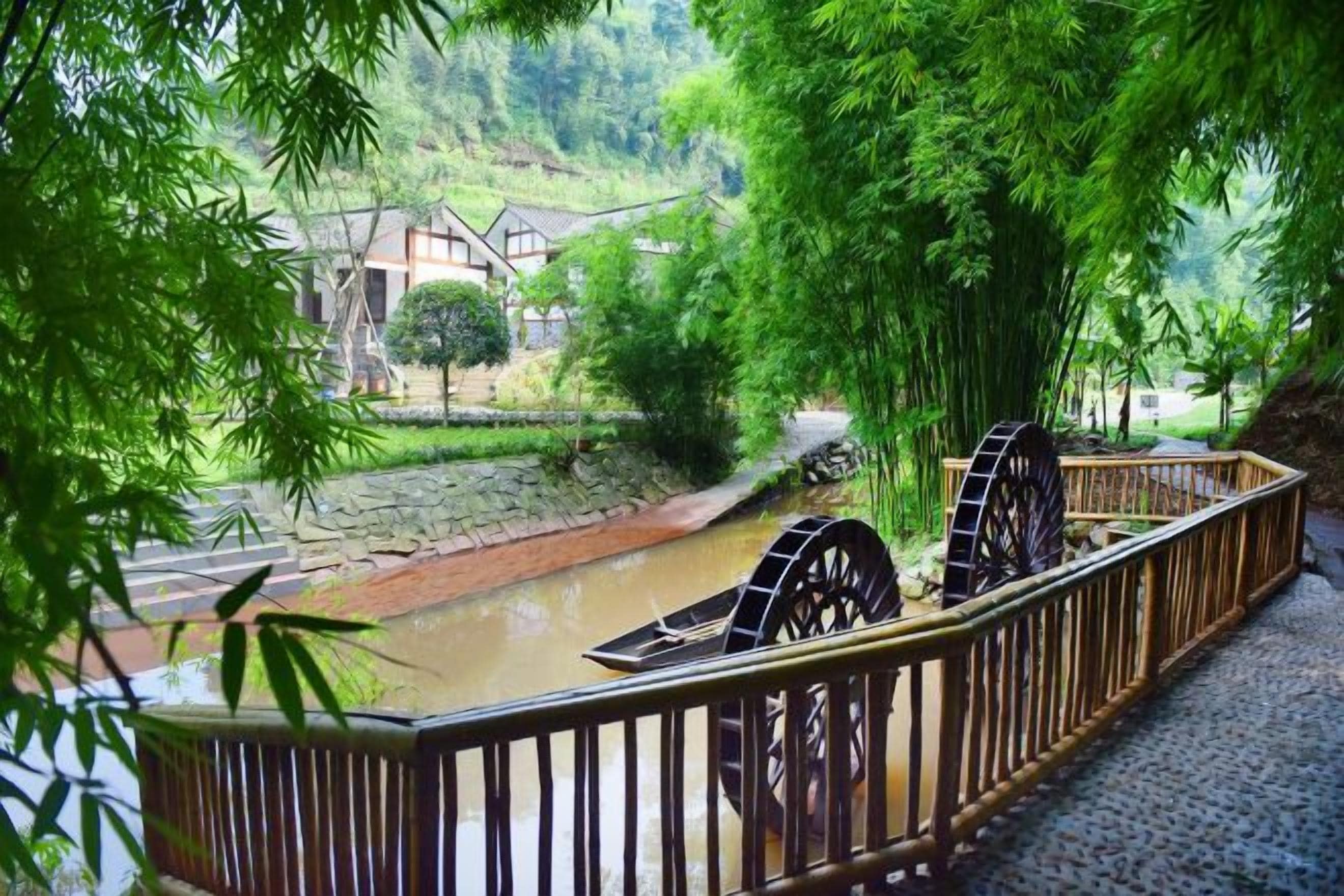 A riverside walkway with a traditional water wheel, bamboo trees, and a stone bridge.