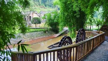 A riverside walkway with a traditional water wheel, bamboo trees, and a stone bridge.