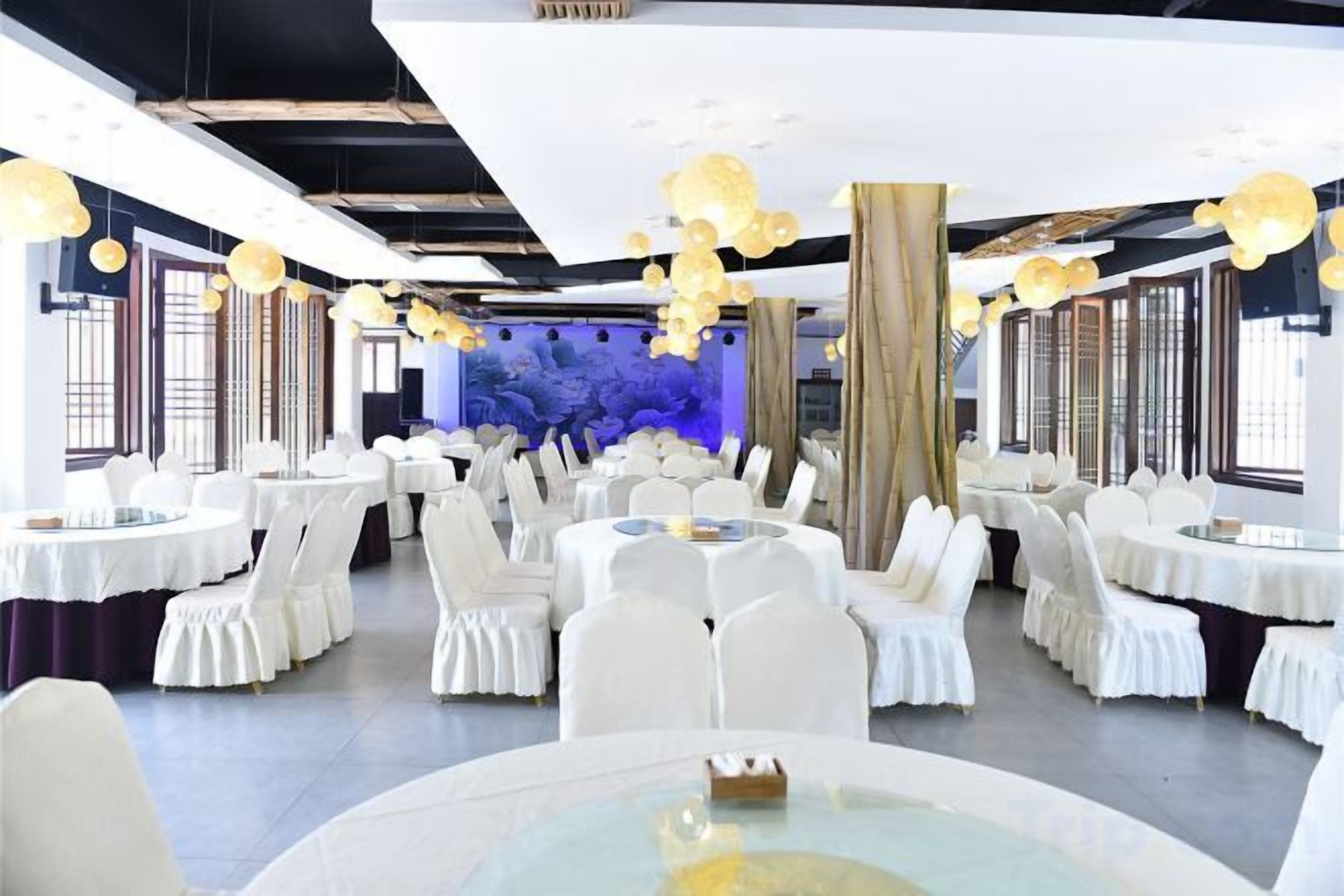 Un salón de banquetes con mesas redondas y sillas blancas, ventanas amplias y lámparas colgantes.
