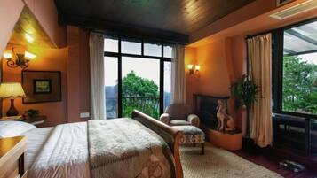 A bedroom with a large bed, a chair, a fireplace, and a view of greenery outside.