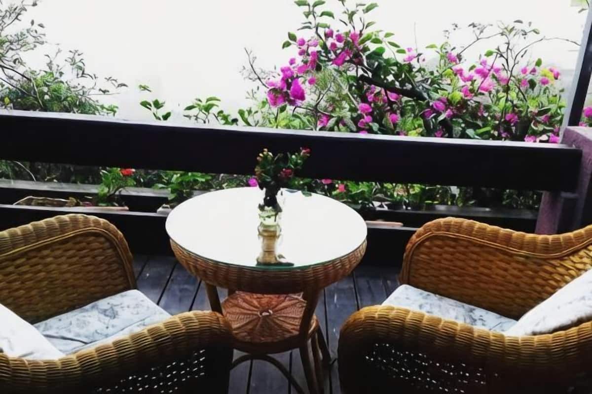 A balcony with wicker chairs and a small round table, surrounded by lush greenery and pink flowers.
