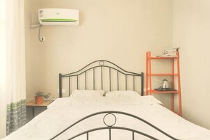 Una cama con estructura metálica, una mesita de noche con una planta, un aire acondicionado instalado en la pared y un estante rojo con libros y una tetera.