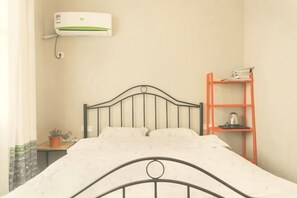 A bed with a metal frame, a small bedside table with a plant, a wall-mounted air conditioner, and a red shelf with books and a kettle.