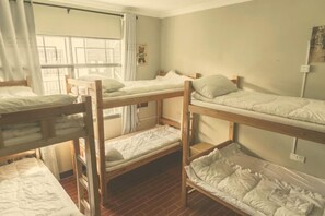 A dormitory room with bunk beds, a window with curtains, and a small bedside table.