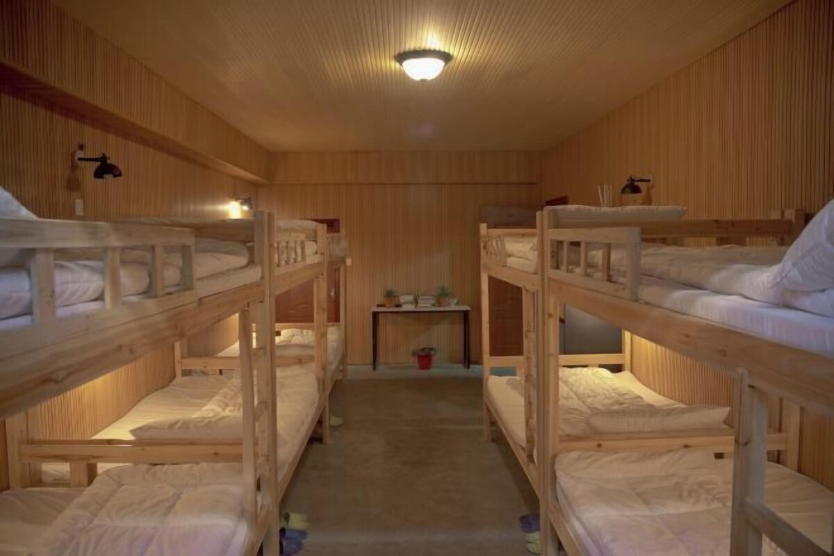 Huoneessa on rivissä kerrossängyt, pieni pöytä, jonka päällä on tavaroita, ja kattovalaisin.