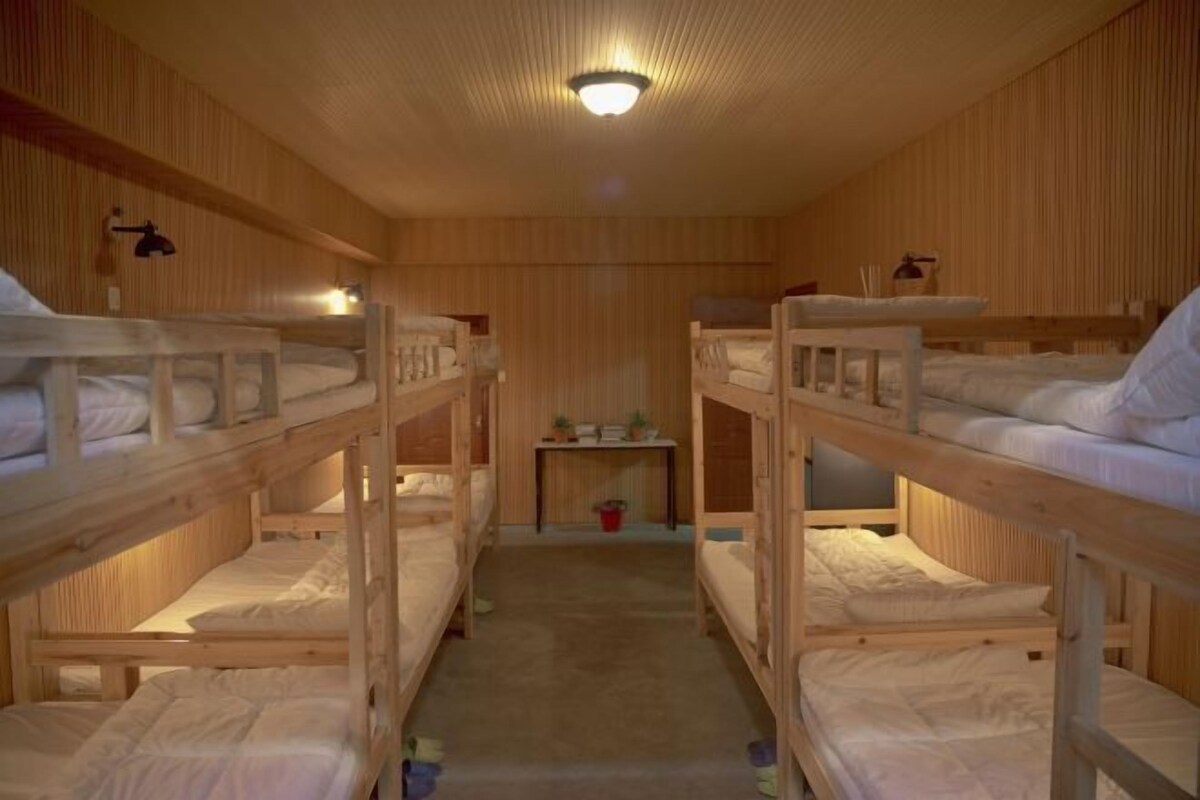 Huoneessa on kerrossängyt, jotka on järjestetty kahteen riviin, pieni pöytä lampun kanssa ja kattovalaisin.