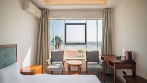 Et hotelværelse med en seng, et skrivebord, to stole, et lille bord og et stort vindue med udsigt over stranden og havet.