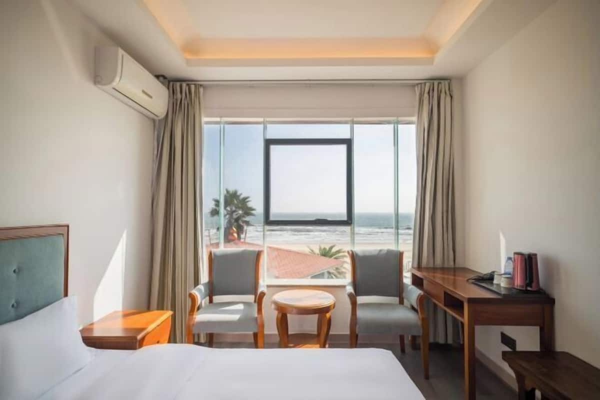 Une chambre d’hôtel comprenant un lit, un bureau, deux chaises, une petite table et une grande fenêtre donnant sur la plage et l’océan.
