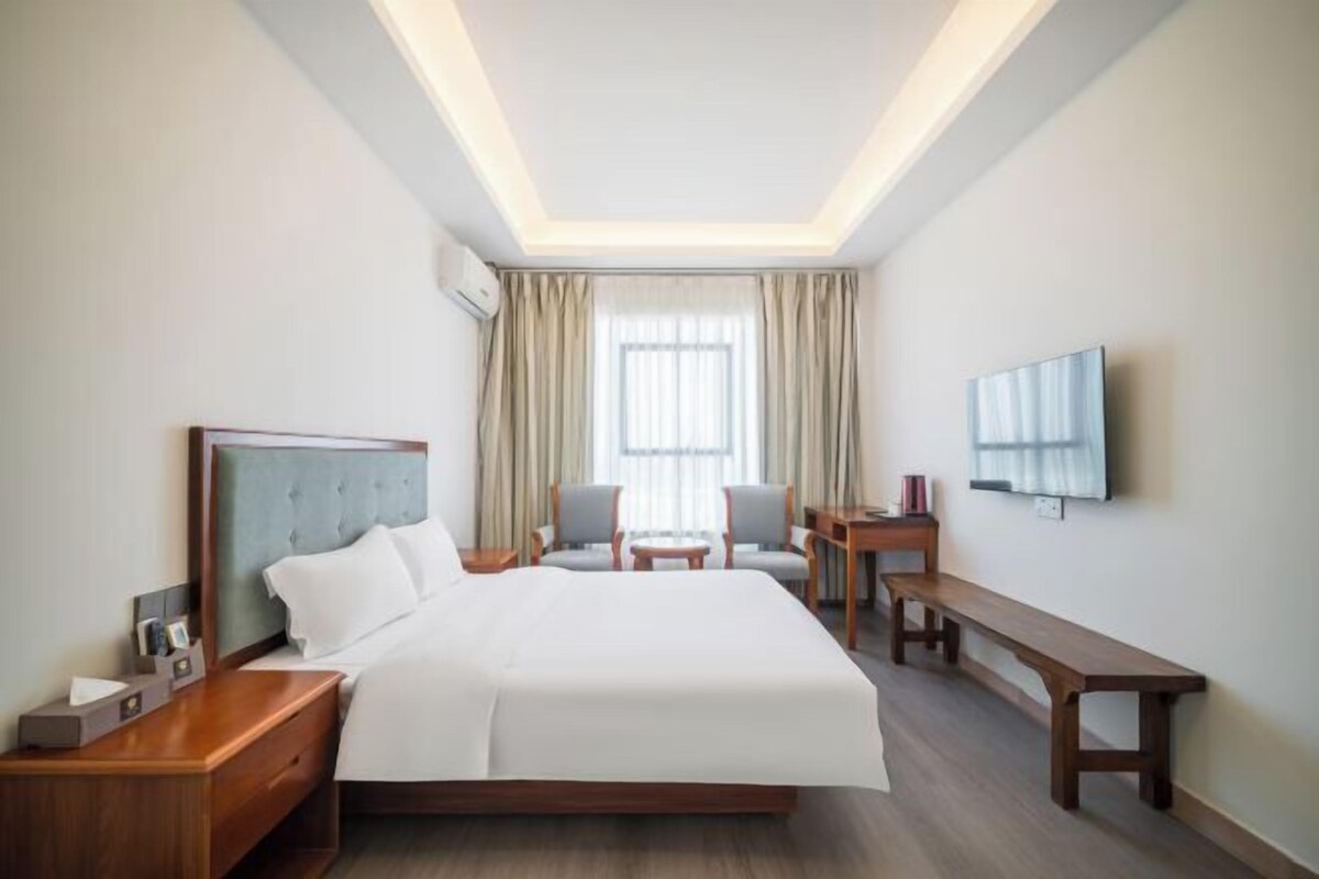 Une chambre d’hôtel avec un grand lit, un bureau, deux chaises, un banc, une télévision et une fenêtre avec des rideaux.