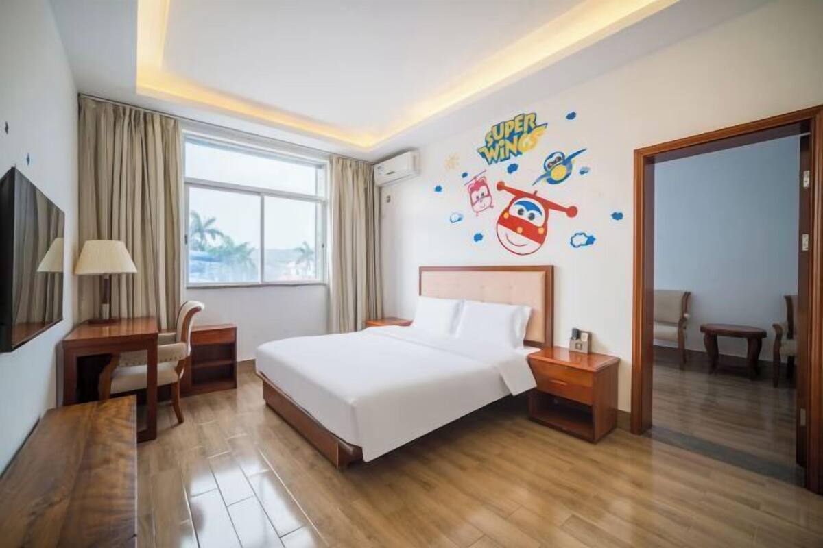 Une chambre d’hôtel avec un grand lit, un bureau, une chaise, une table de chevet et un mur orné d’une fresque représentant les personnages de Super Wings.