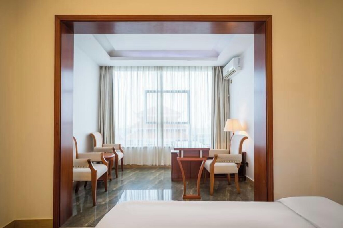 Une chambre d’hôtel comprenant un lit, deux fauteuils, une petite table, une fenêtre avec des rideaux et un appareil de climatisation.
