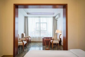 Una habitación de hotel con una cama, dos sillones, una mesita, una ventana con cortinas y un equipo de aire acondicionado.
