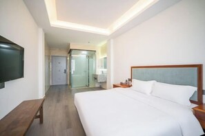 A hotel room with a large bed, a wooden bench, a flat-screen TV, and a glass shower enclosure. - Xinsha Hotel (Zhanjiang)