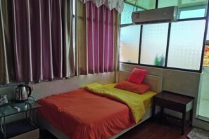Una cama individual con colcha roja y amarilla, una mesita de noche pequeña y una ventana con cortinas.
