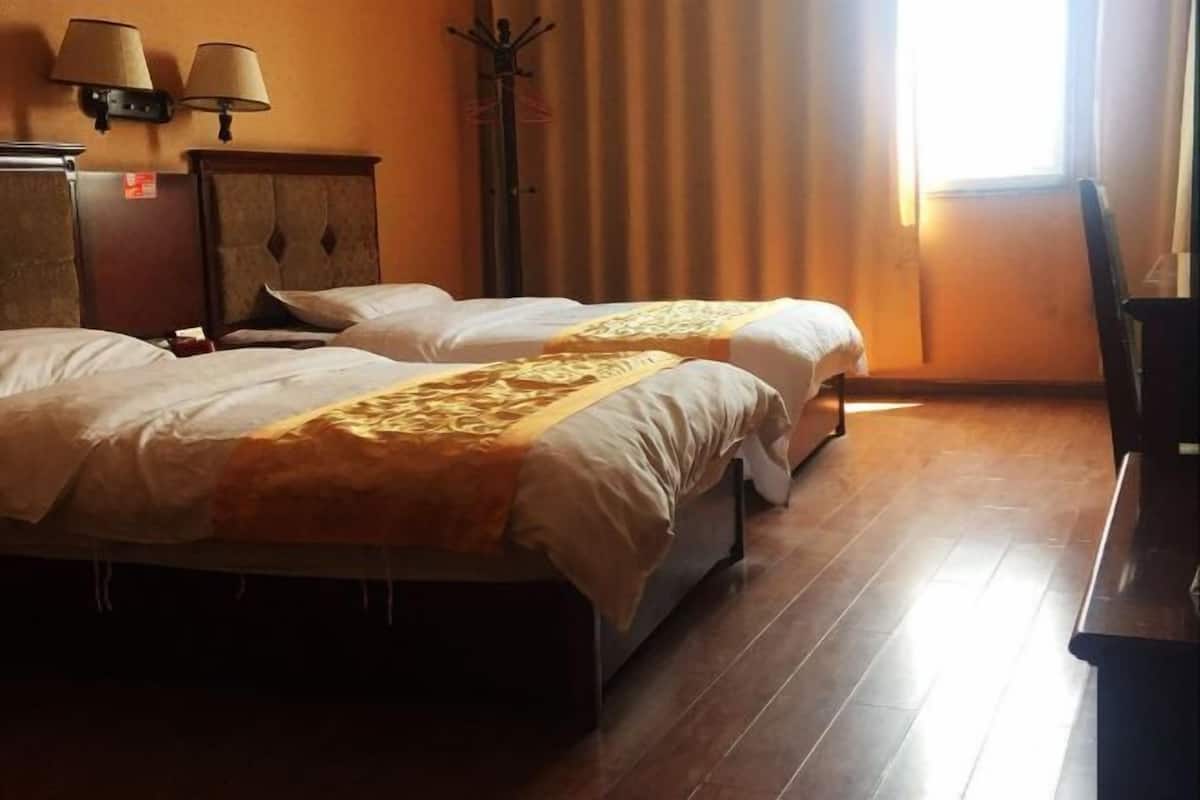 A hotel room with two beds, a wooden floor, a nightstand, a lamp, and a window with curtains.