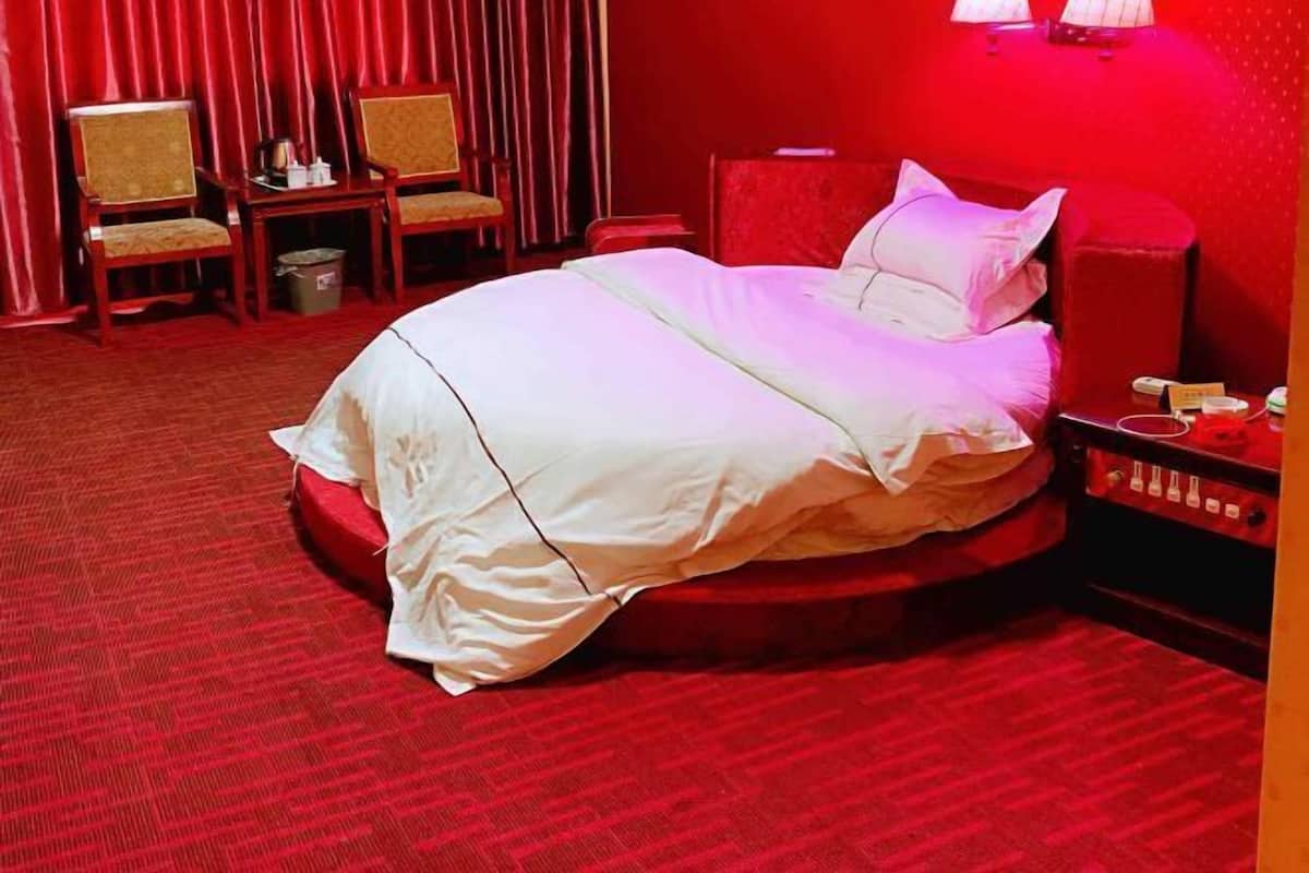 A hotel room with a red carpet, a bed with white linens, two wooden chairs, a small table with a lamp, and a nightstand with a phone and a book.