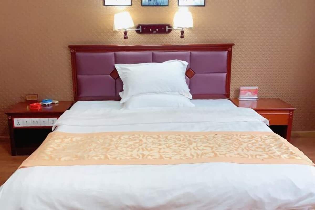 A hotel room with a large bed, a wooden headboard, two bedside lamps, and a nightstand with a lamp and a red object.